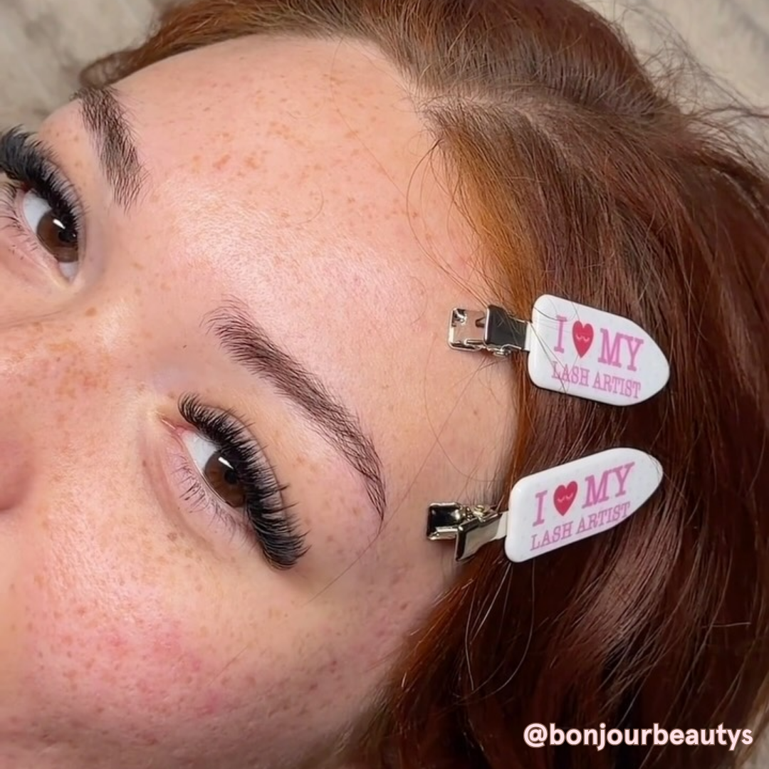 Close-up of a lash client with full eyelash extensions wearing two “I Love My Lash Artist” hair clips, with auburn hair and freckles visible.