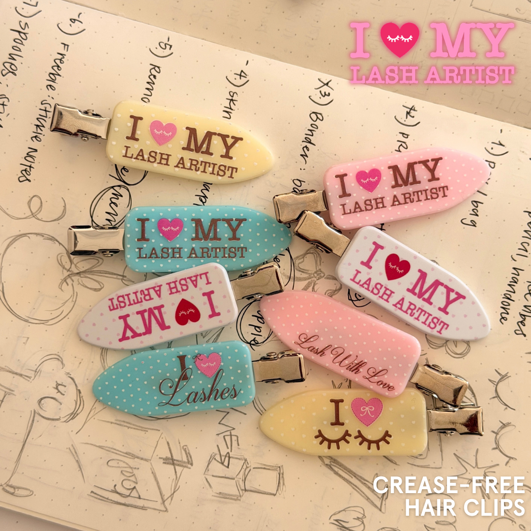 Colorful crease-free hair clips featuring “I Love My Lash Artist” and lash-themed designs in pink, blue, yellow, and white, arranged over a sketchbook.