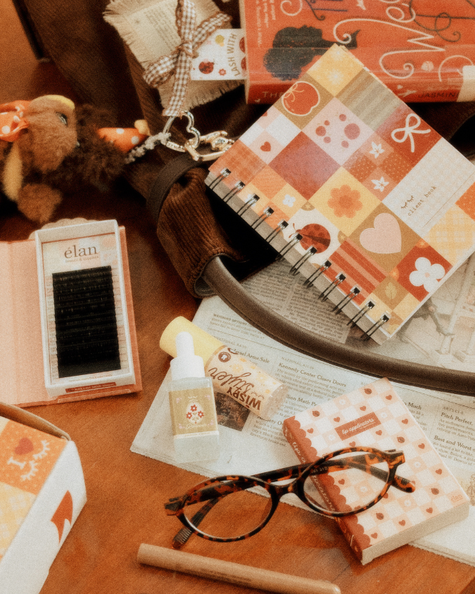 Book-themed flat lay featuring a client book, lash tray, Wispy Styler, protein remover, lip applicators, glasses, and cozy reading accessories in warm orange and brown tones.