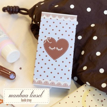 Manhua Tinsel Lash Tray with brown polka dots and light blue packaging with a brown heart and eyelashes illustrated on it laying on a brown and white polka dot bag.
