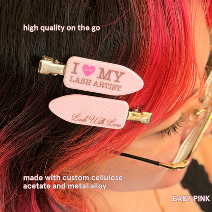 Baby pink “I Love My Lash Artist” crease-free hair clips worn in black and red hair, with text highlighting durable cellulose acetate and metal alloy construction.