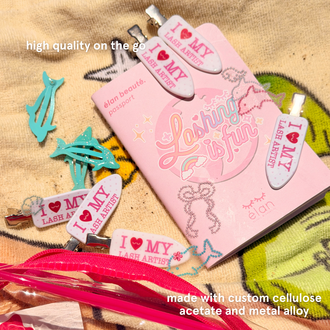 Crease-free hair clips with text on it saying "I Heart My Lash Artist" in pink text laying on a beach towel with text saying "High quality on the go" and "Made with custom cellulose acetate and metal alloy"