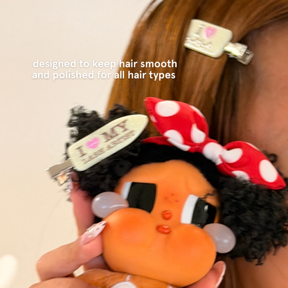 Close-up of cream “I Love My Lash Artist” hair clips securing smooth hair, with a doll held in the foreground and text highlighting a crease-free design for all hair types.