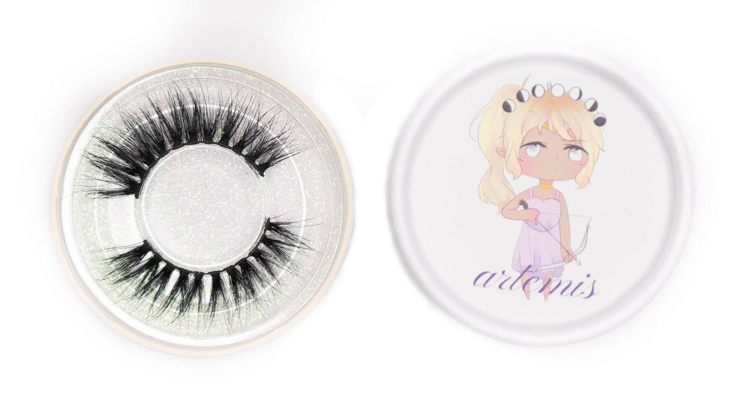 Artemis false eyelashes in a clear round case beside the illustrated Artemis product lid on a white background.