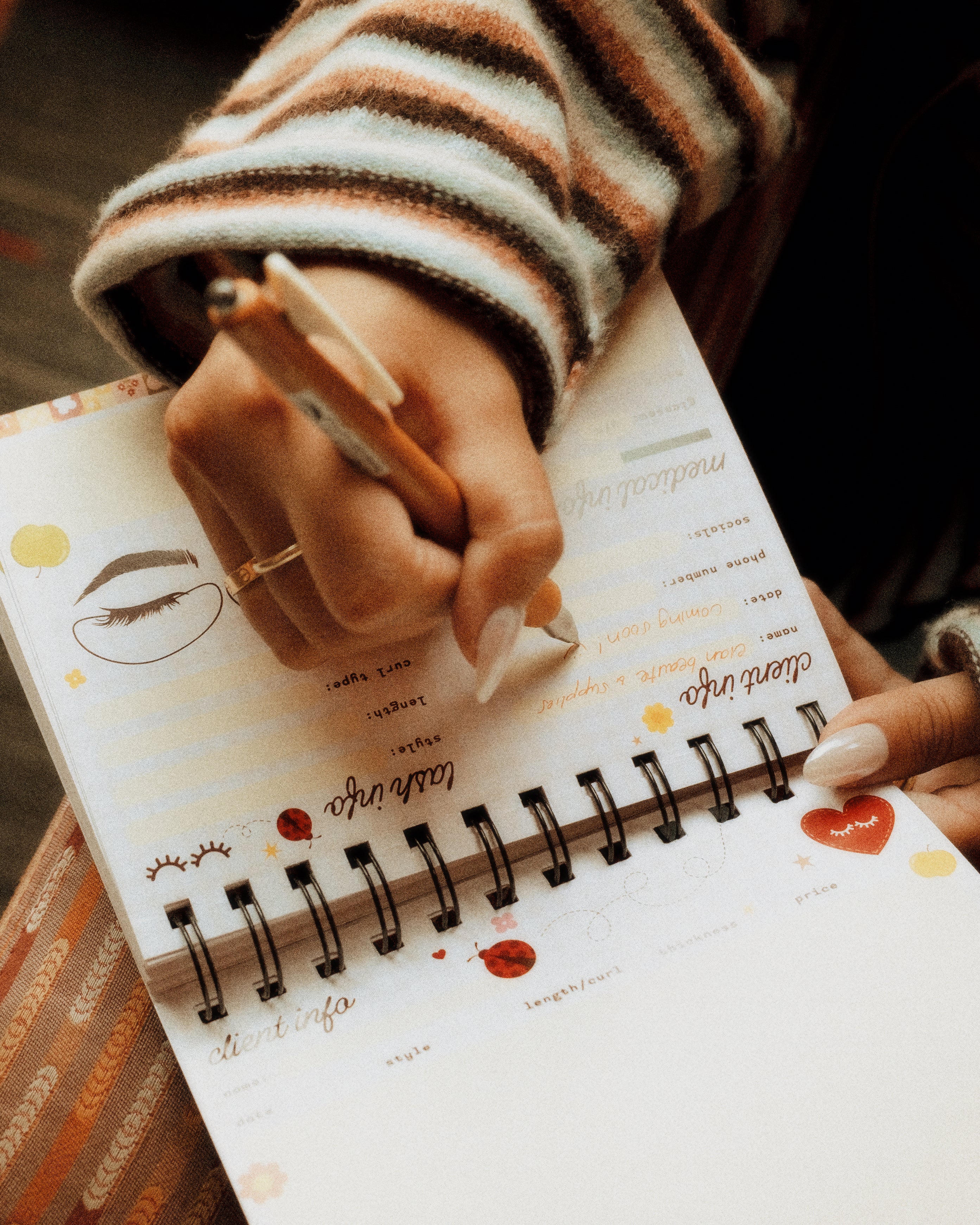 Close-up of a person writing in a spiral-bound lash client book featuring client information and lash mapping sections, with warm floral and ladybug details.