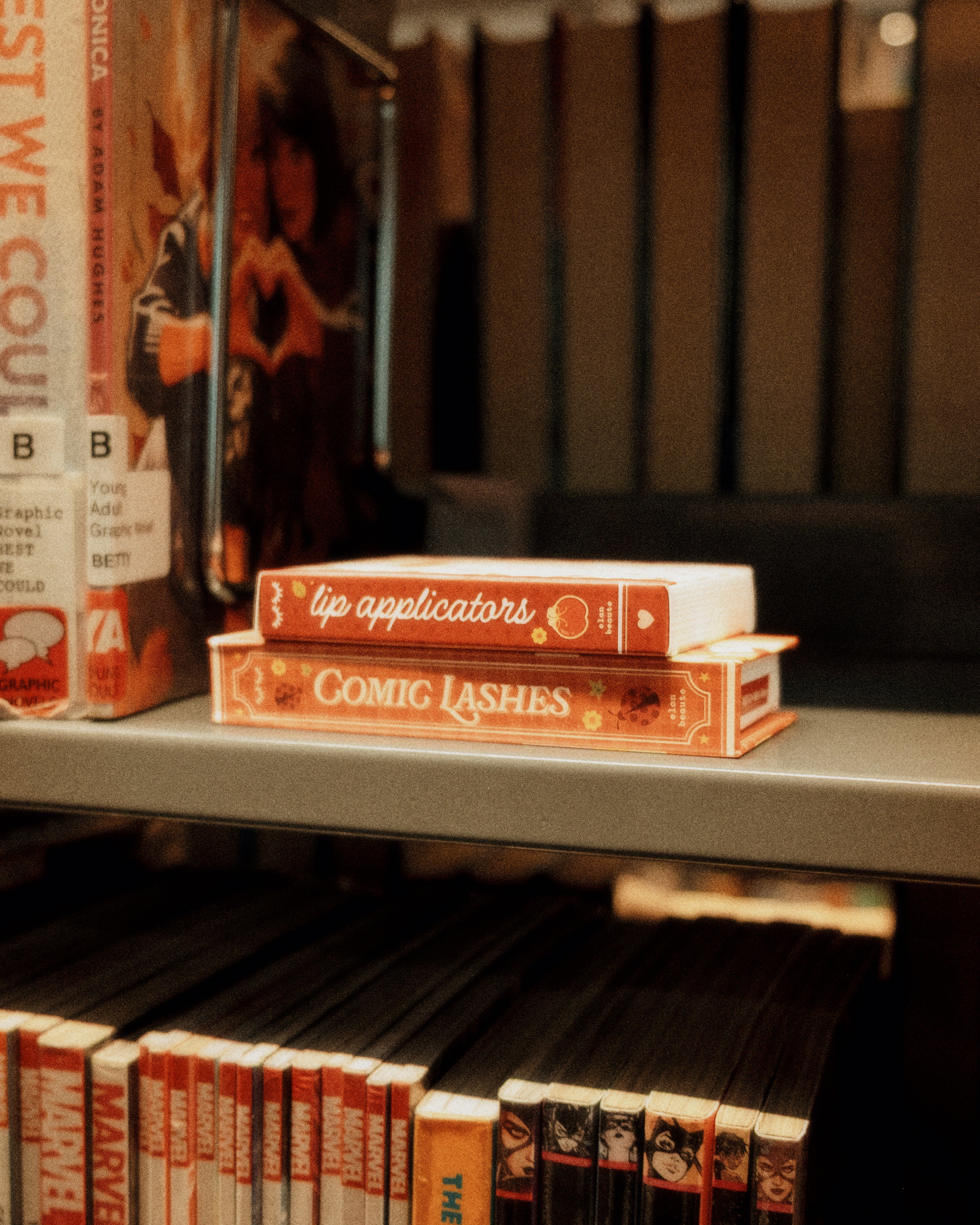 Comic Lashes and lip applicator boxes styled like books on a library shelf, featuring warm red and orange vintage-inspired packaging.