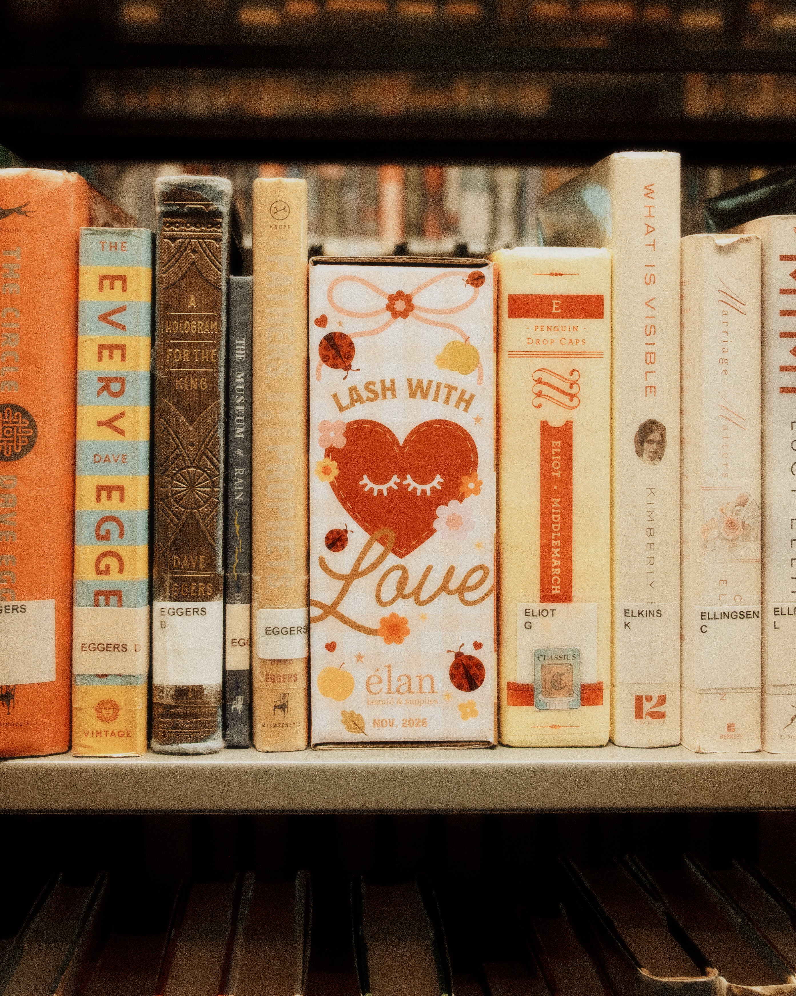 Book-themed “Lash With Love” Élan Beauté November 2026 box displayed upright on a library shelf between books, featuring warm floral, heart, and ladybug graphics.