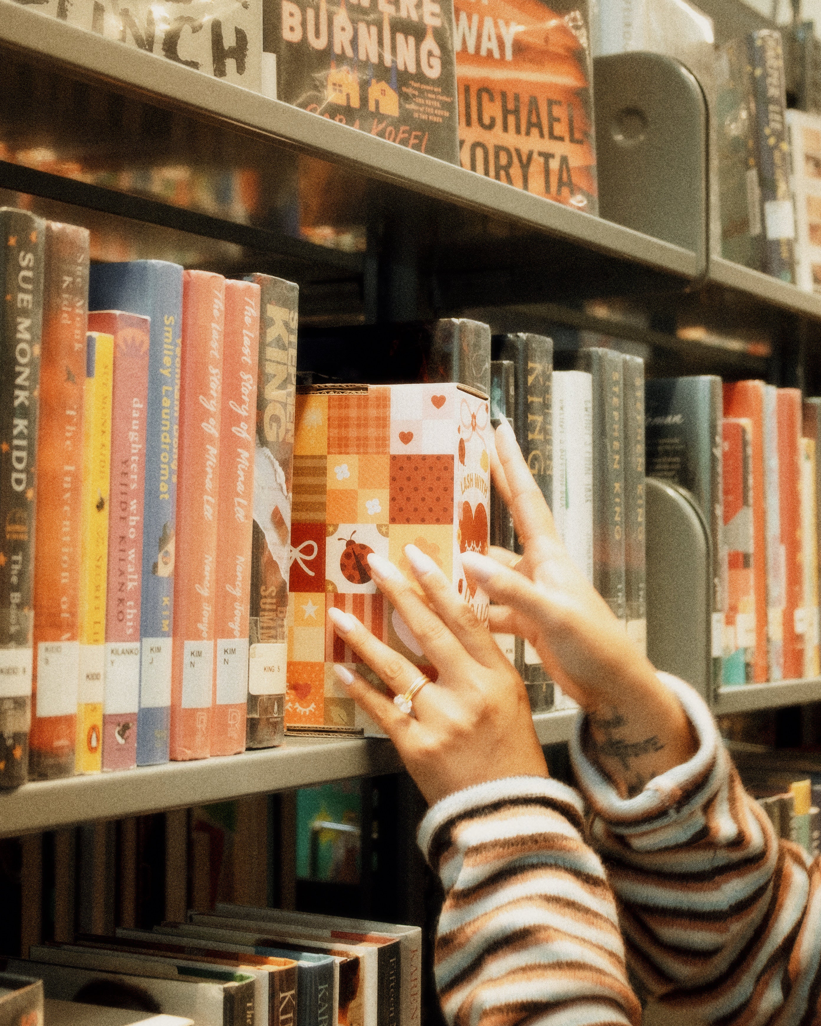 Book-themed Élan Beauté subscription box being placed on a library shelf among books, featuring warm patchwork packaging with hearts, flowers, and ladybug details