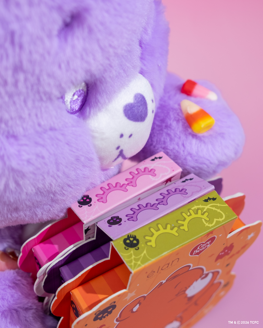 Élan Beauté x Care Bears Big Boi lash trays featuring pink, orange, and purple Halloween-themed character packaging displayed beside a purple Care Bear plush on a pink background.