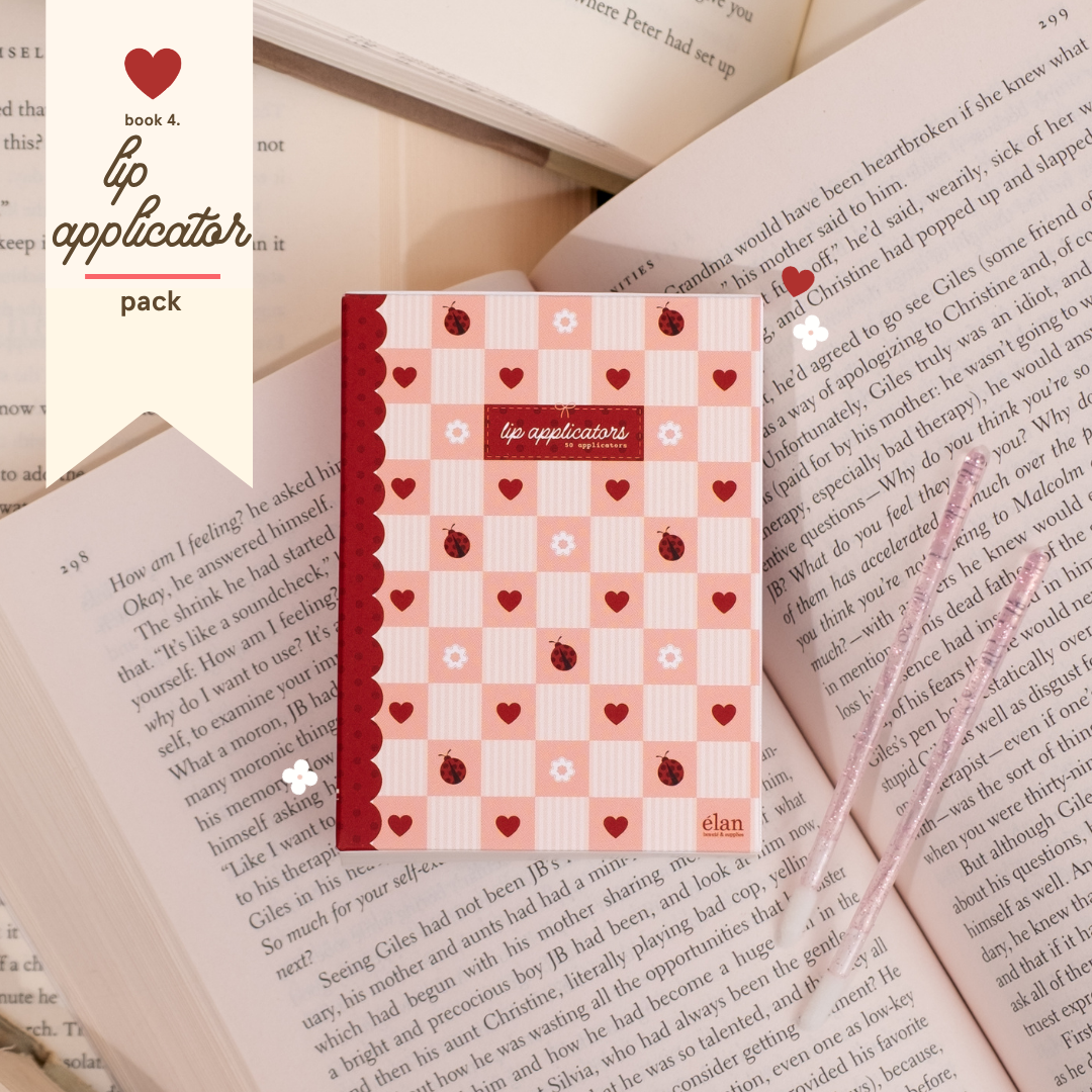 Lip applicator pack in pink and red heart-patterned packaging, styled on top of open book pages with two glittery applicator wands beside it.