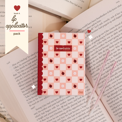 Lip applicator pack in pink and red heart-patterned packaging, styled on top of open book pages with two glittery applicator wands beside it.
