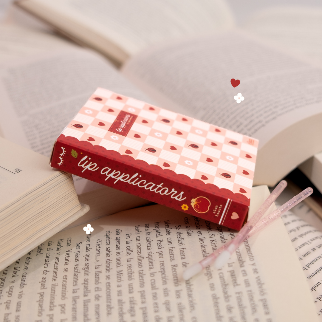 Pink and red lip applicator box with two glittery applicator wands, styled across open book pages with small heart and flower accents.