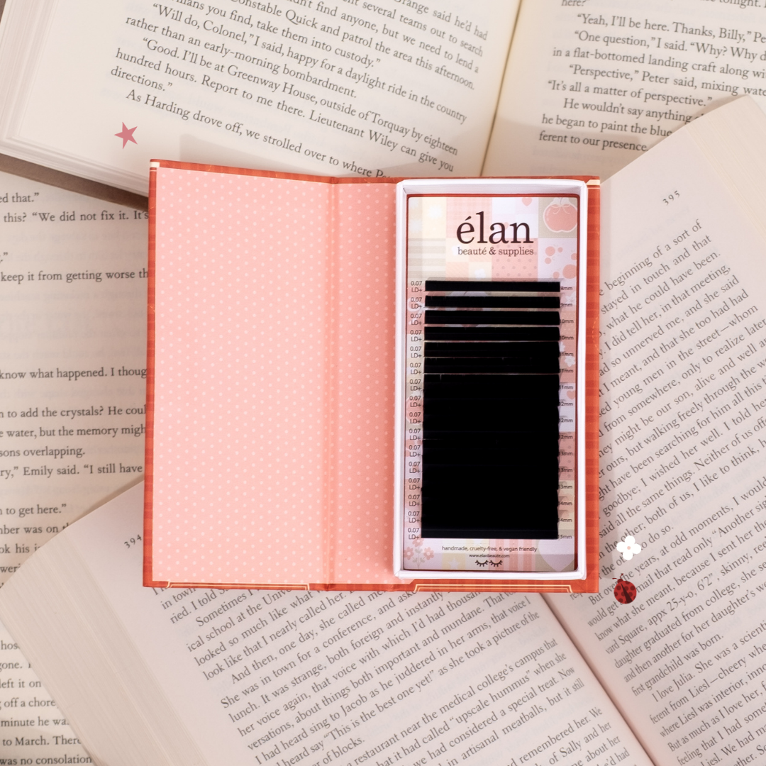 Open Comic Lashes tray displayed like a book, featuring black lash extensions inside red and pink packaging, styled on top of open book pages.
