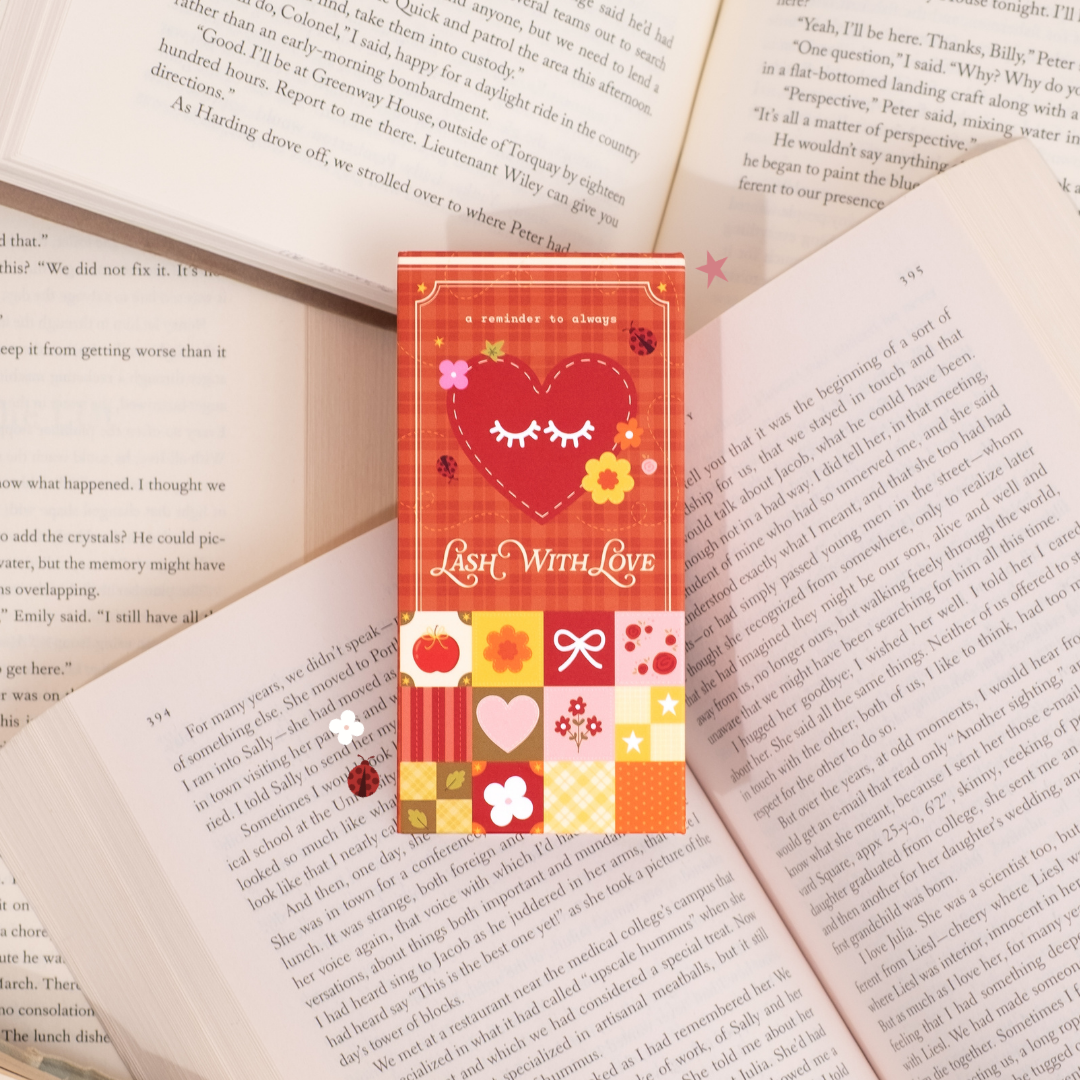 Red and orange “Lash With Love” packaging featuring a heart with eyelash graphics, floral details, and a patchwork pattern, styled on top of open book pages.