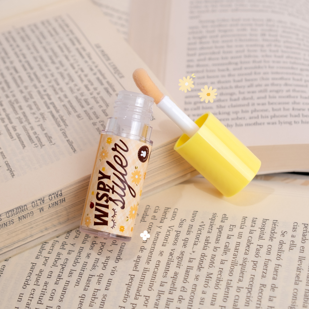 Wispy Styler applicator shown open with its soft wand, styled on top of open book pages with yellow floral accents.