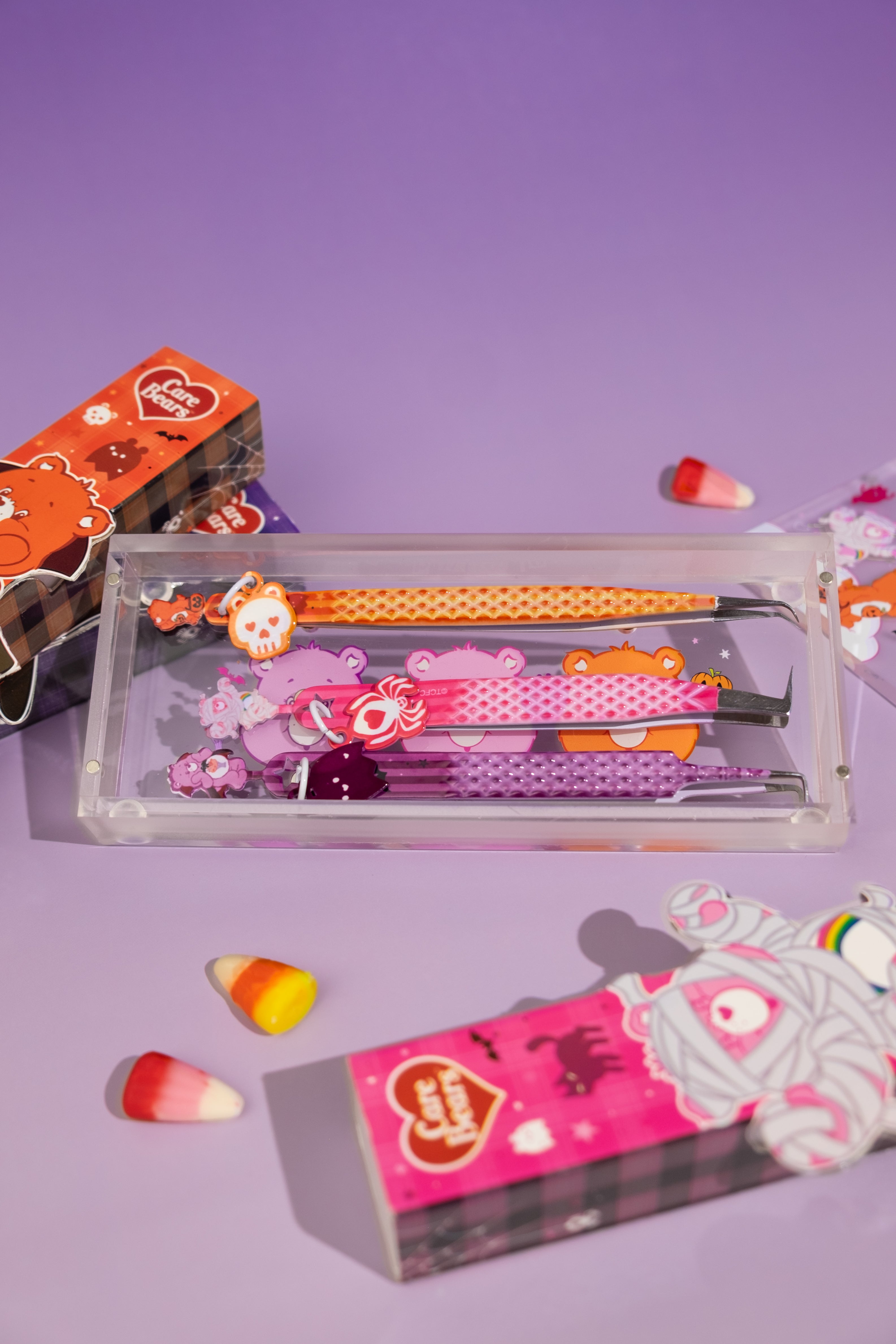 Élan Beauté x Care Bears Halloween tweezer set featuring orange, pink, and purple lash tweezers with character charms displayed in a clear magnetic case, surrounded by matching Care Bears packaging and candy corn accents.