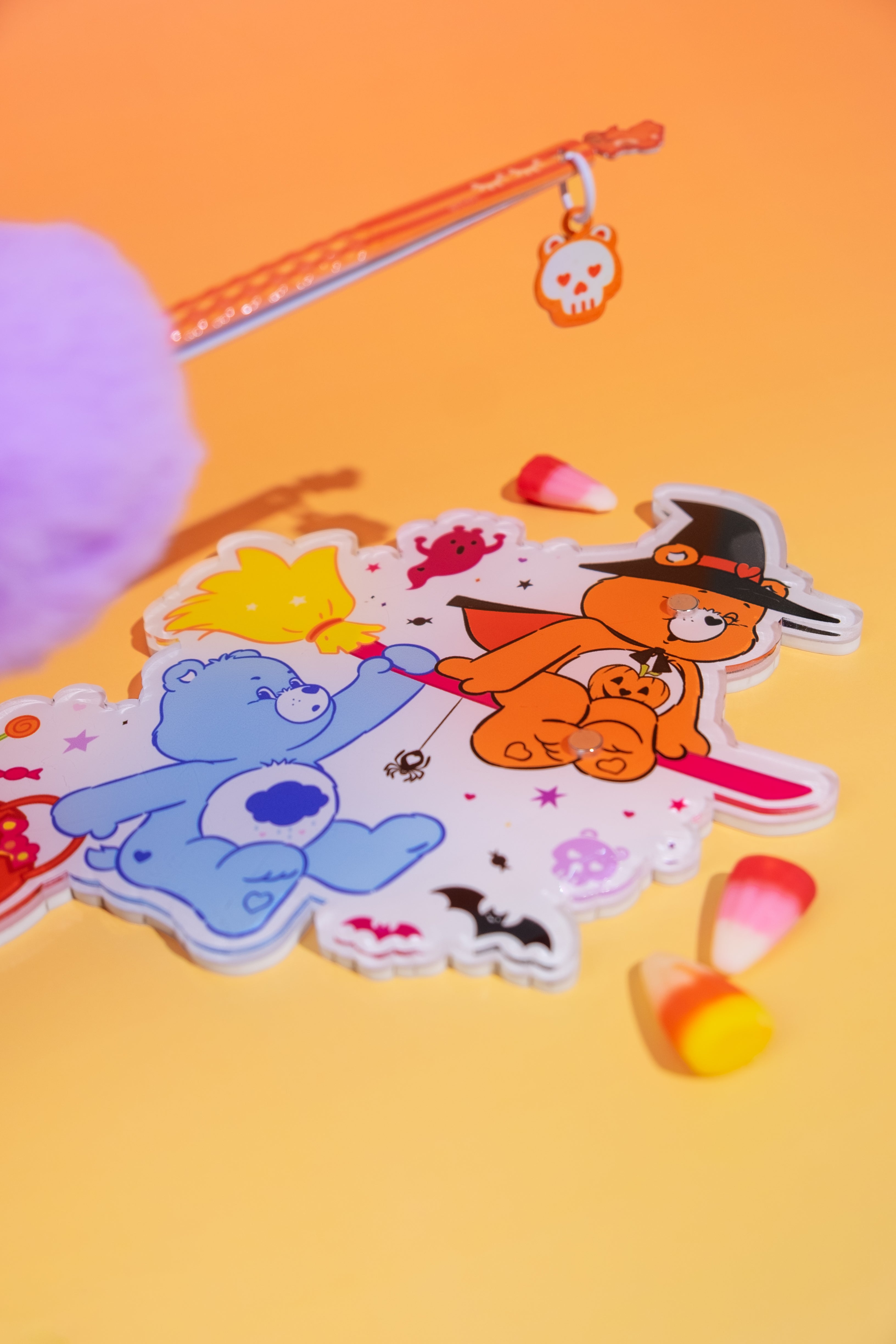 Élan Beauté x Care Bears Halloween lash tile featuring orange and blue Care Bears on a witch’s broom, styled with an orange skull-charm tweezer and candy corn on a warm orange background.