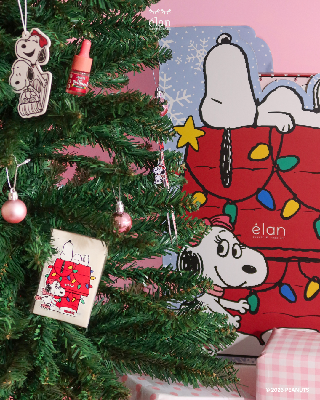 PEANUTS holiday display featuring a Christmas tree decorated with Snoopy and Belle ornaments, Élan Beauté products, pink baubles, and festive Snoopy packaging in the background.