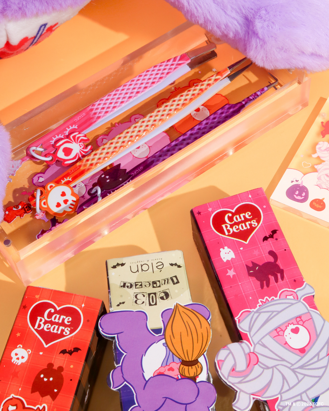 Élan Beauté x Care Bears Halloween collection featuring pink, orange, and purple lash tweezers with character charms, matching packaging, and a clear magnetic tweezer case styled on an orange background.