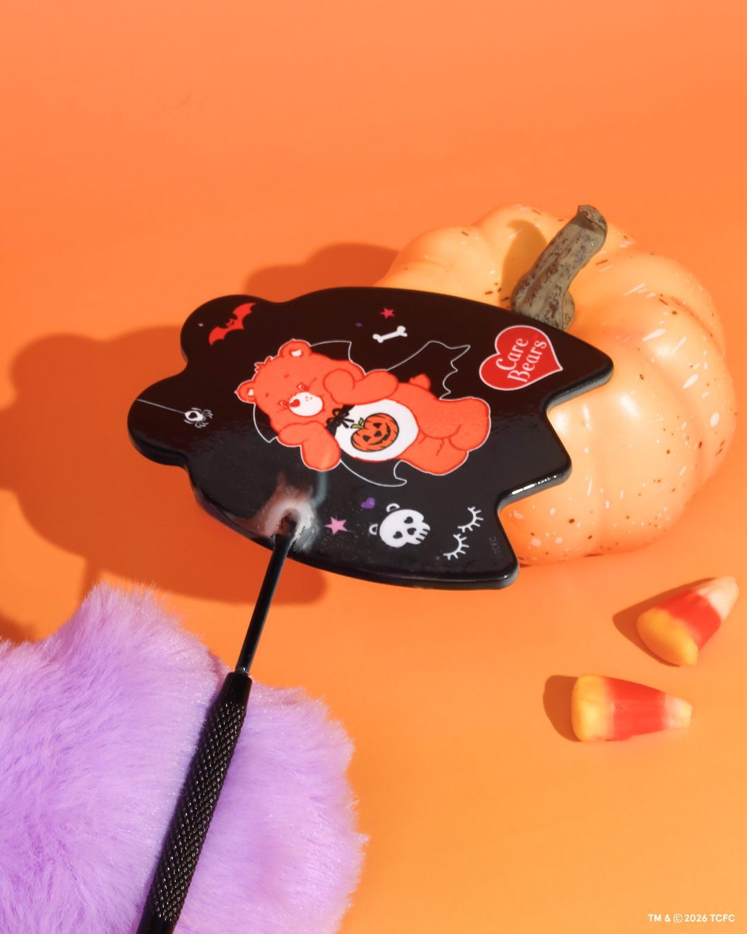 Élan Beauté x Care Bears Halloween handheld mirror featuring an orange Care Bear in a vampire costume with pumpkin, skull, bat, and spider details, styled beside a white pumpkin and candy corn on an orange background.
