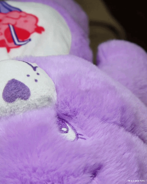 Élan Beauté x Care Bears handheld mirror reflecting a purple Care Bear plush, styled with additional purple Care Bears plush toys in the background.