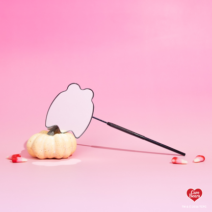 Élan Beauté x Care Bears Halloween handheld mirror with a black textured handle, styled beside a white pumpkin and candy corn on a pink background.