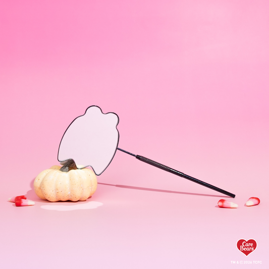 Élan Beauté x Care Bears Halloween handheld mirror with a black textured handle, styled beside a white pumpkin and candy corn on a pink background.