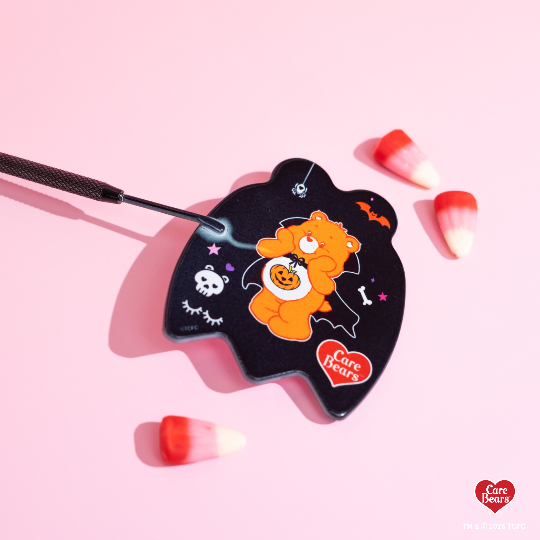 Élan Beauté x Care Bears Halloween handheld mirror featuring an orange Care Bear in a vampire costume with pumpkin, bat, skull, and spider details, styled with candy corn on a pink background.