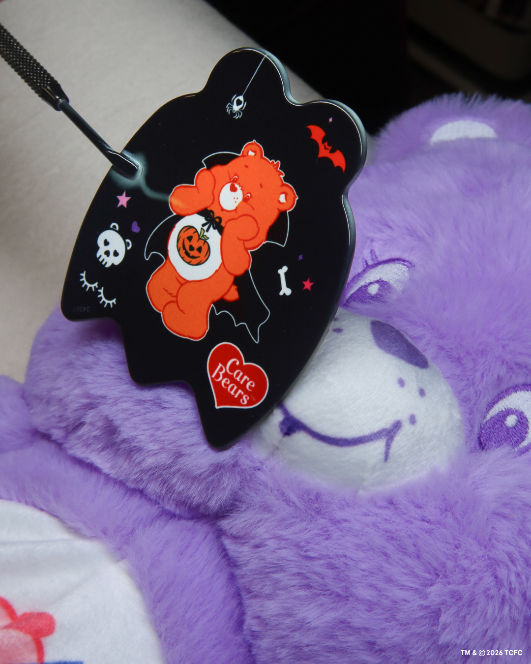 Élan Beauté x Care Bears Halloween handheld mirror featuring an orange vampire Care Bear with pumpkin, bat, skull, and spider details, resting on a purple Care Bear plush.