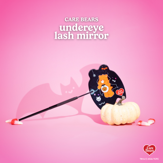 Élan Beauté x Care Bears Halloween undereye handheld lash mirror featuring an orange Care Bear in a vampire costume with pumpkin, bat, skull, and spider details, styled with candy corn on a pink background.