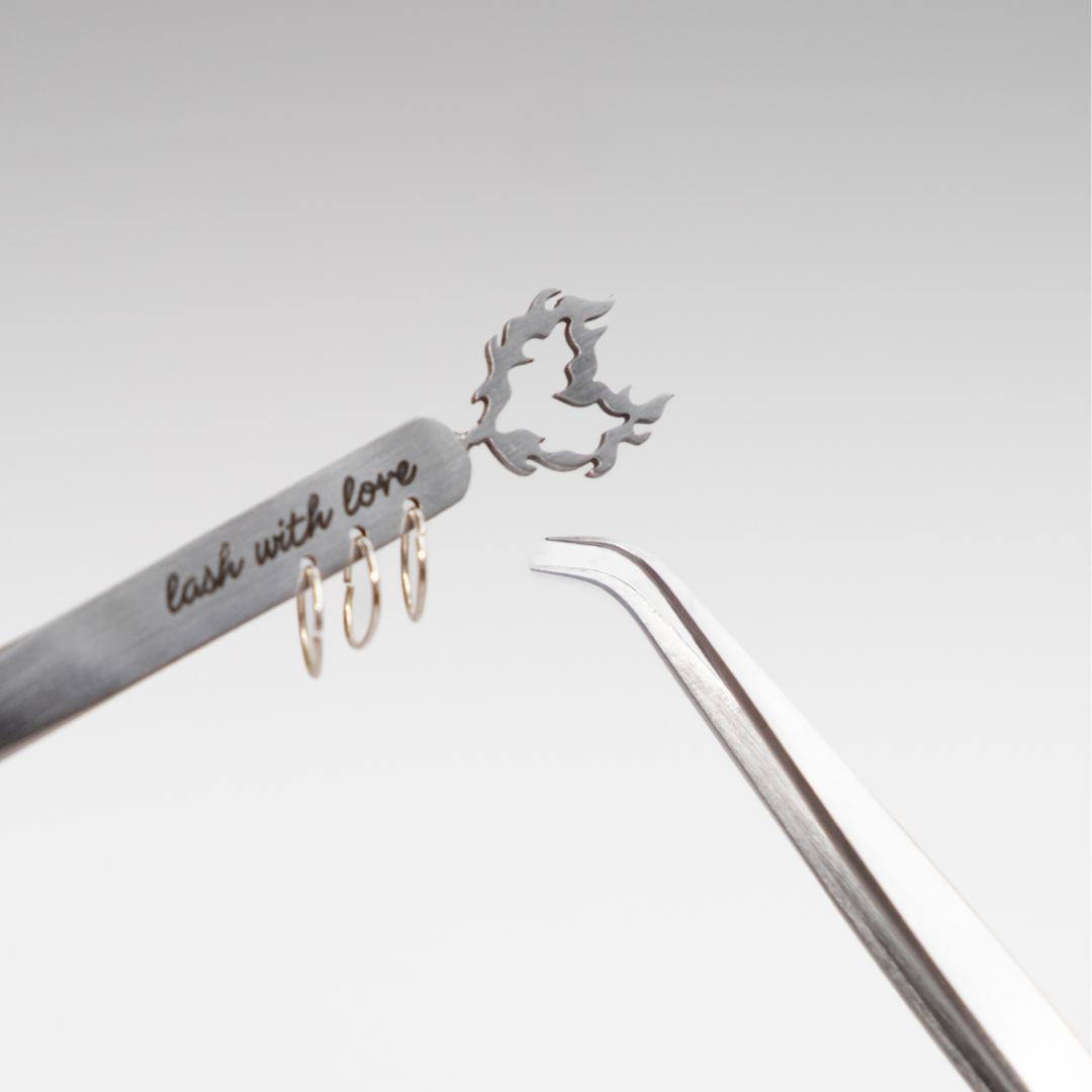 Close-up of stainless steel lash tweezers featuring a curved isolation tip, engraved “lash with love” detail, decorative gold rings, and a flame-shaped cutout.