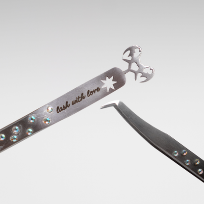 Close-up of stainless steel lash tweezers featuring a curved isolation tip, engraved “lash with love” detail, rhinestone accents, and a heart-shaped bat-wing cutout.