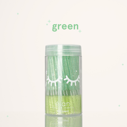 Élan Beauté green glitter spoolies in a clear 100-count container with white eyelash graphics, displayed on a soft cream background with green accents.