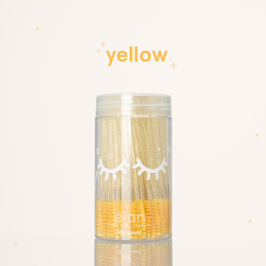 Élan Beauté yellow glitter spoolies in a clear 100-count container with white eyelash graphics, displayed on a soft cream background with yellow accents.