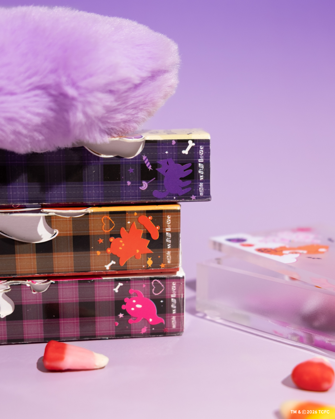 Élan Beauté x Care Bears Halloween collection featuring stacked purple, orange, and pink lash tray boxes, a clear magnetic case, purple Care Bear plush paw, and candy corn accents on a lavender background.